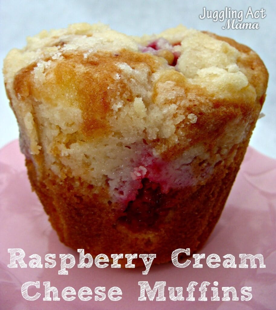 A close-up of a raspberry cream cheese muffin with a crumbly top, showing bits of raspberry, on a pink surface. The text reads Raspberry Cream Cheese Muffins and Juggling Act Mama.
