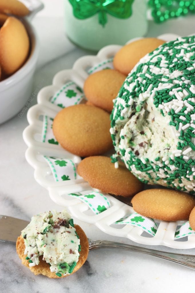 A mint cheeseball covered with green and white sprinkles in the back ground and a vanilla wafer cookie with some of the mint cheese spread on it.