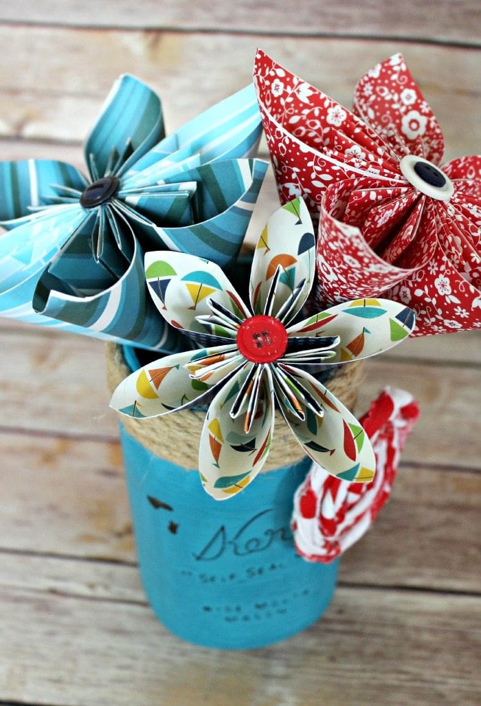 Red, white and blue paper flowers made with patterned paper in a blue mason jar with jute cording around the top and a white and red rolled fabric rose.