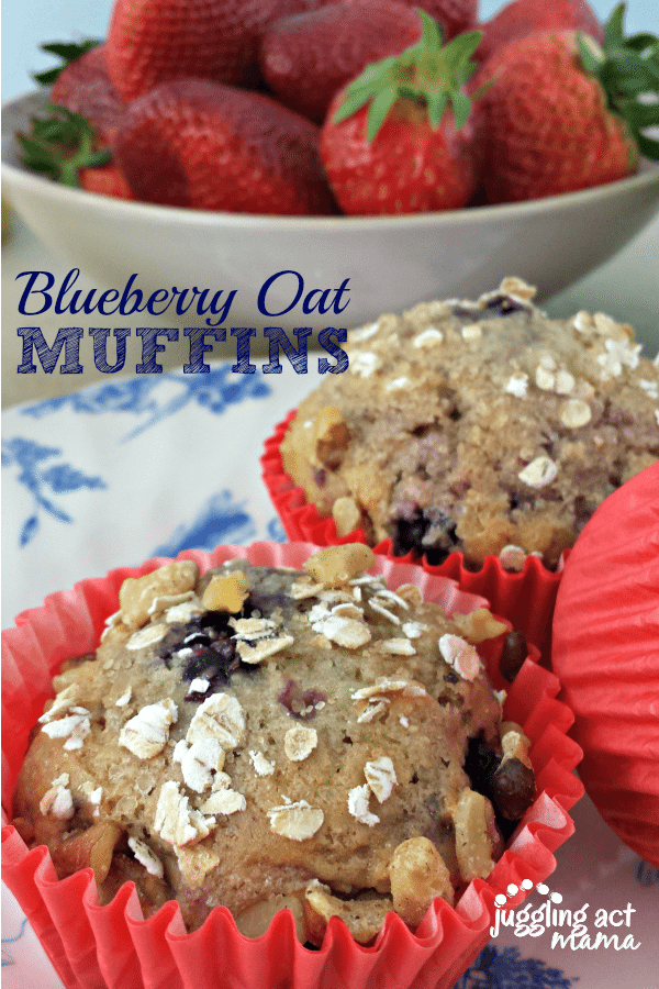 2 blueberry oat muffins wrapped in a red cupcake wrapper with a white bowl of strawberries in the distance with the text "blueberry oat muffins."