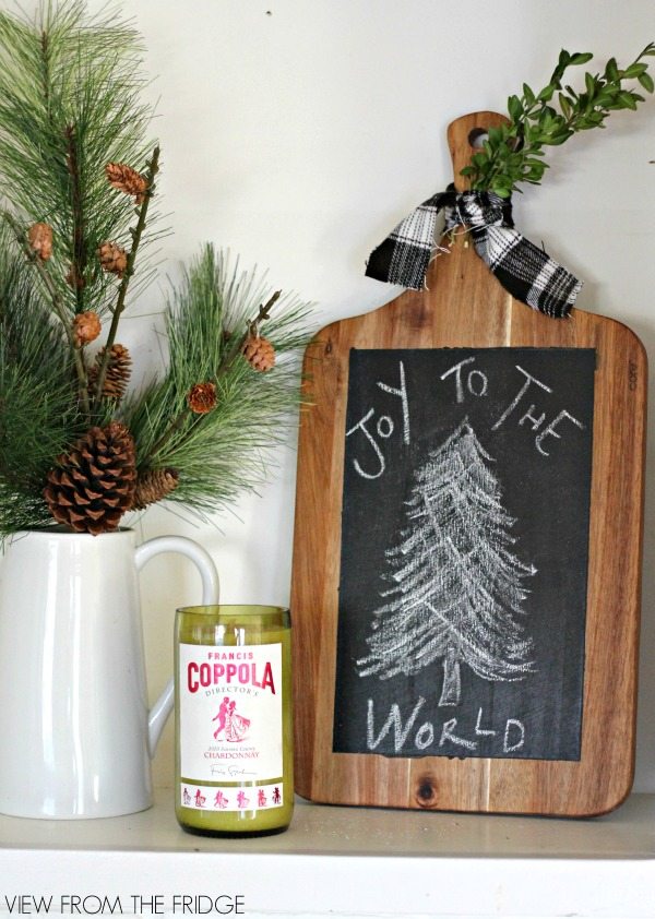 Joy to the World Cutting Board Sign next to a pitcher and candle with Christmas greenery.
