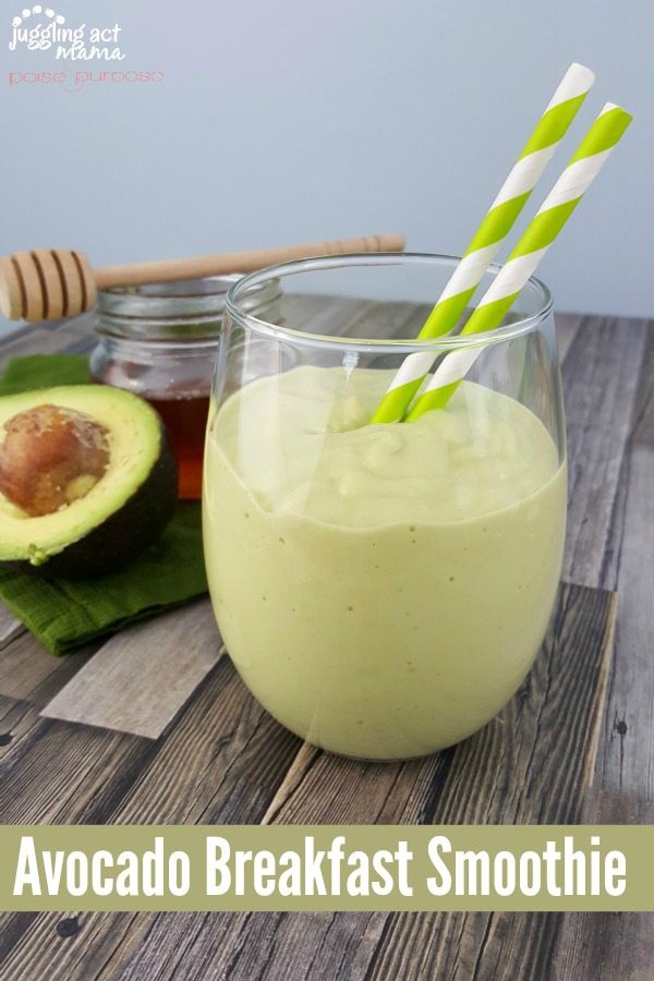 An This avocado and banana smoothie in a small glass with green and white stripped straws next to a halved avocado on a wooden surface.