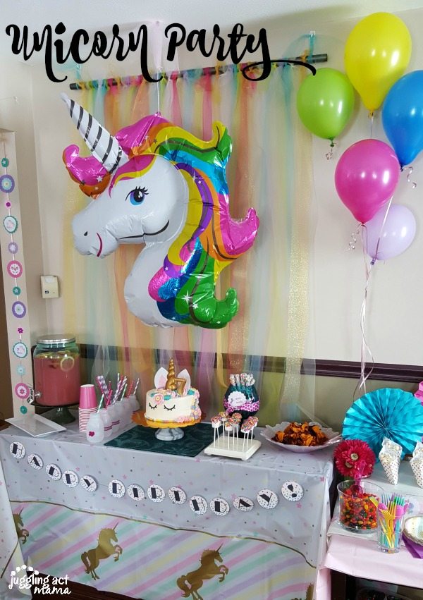 a dessert table at a unicorn party featuring a giant unicorn balloon, streamers pink lemonade, marshmallow pops, a unicorn cake, and other decorations