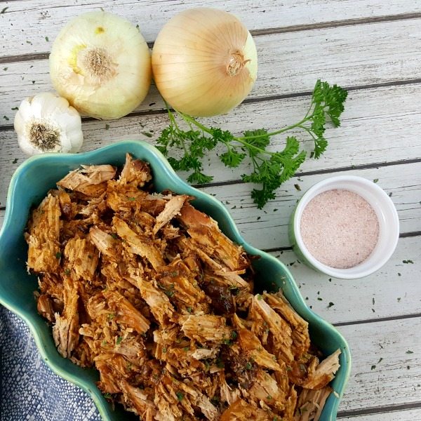 Make Ahead Slow Cooker Pulled Pork in a blue bowl, surrounded by onions, garlic and seasonings.
