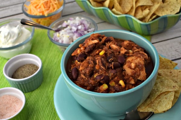Slow Cooker Chicken Chili in a blue bowl on top of a plate filled with tortilla chips and surrounded by topping suggestions - onions, cheese, sour cream.