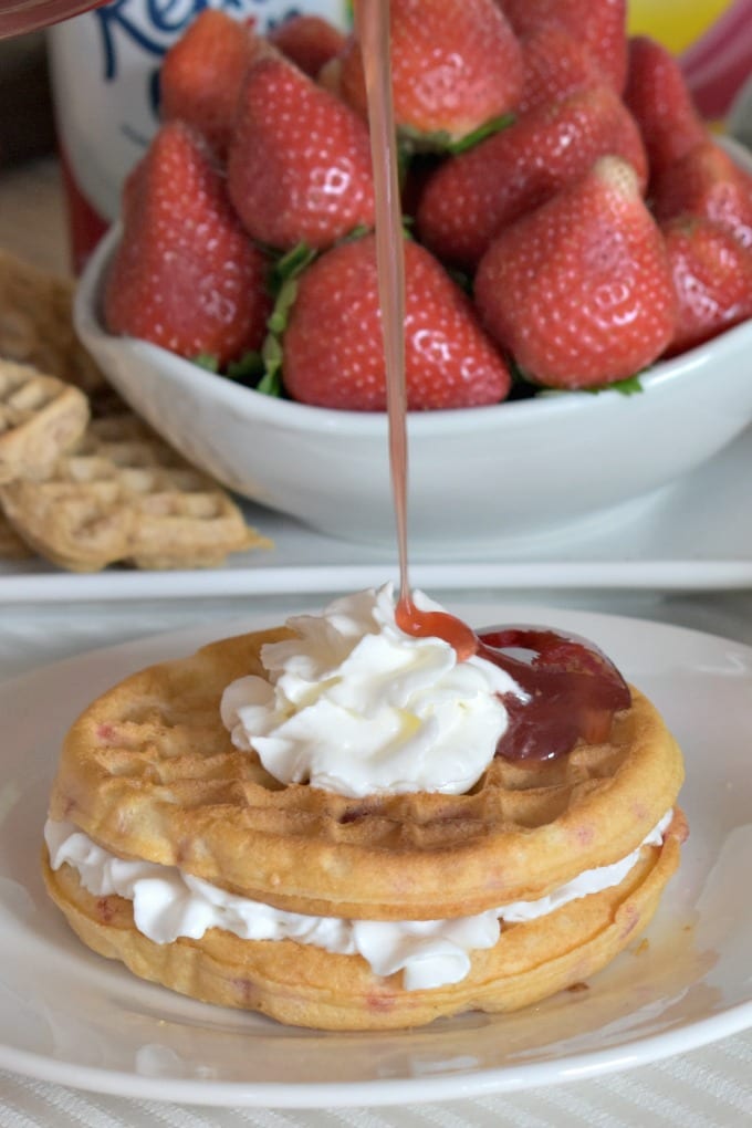 A drizzle of strawberry syrup falls onto strawberry waffles with whipped cream