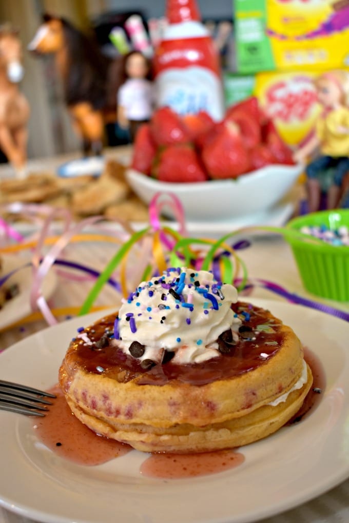 two waffles topped with strawberry syrup and whipped creeam, on a table set with party decorations and waffle toppings.