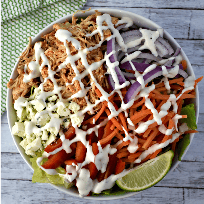 A bowl of salad with shredded chicken, sliced red onions, shredded carrots, diced red bell pepper, crumbled cheese, and a lime wedge, all drizzled with ranch dressing. The bowl sits on a wooden surface.