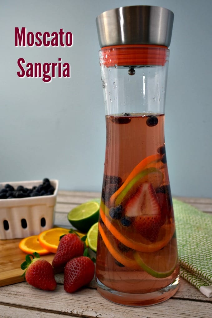 A ceramic berry basket with blueberries, a small cutting board with sliced oranges, limes and strawberries and a carafe with pink moscato sangria