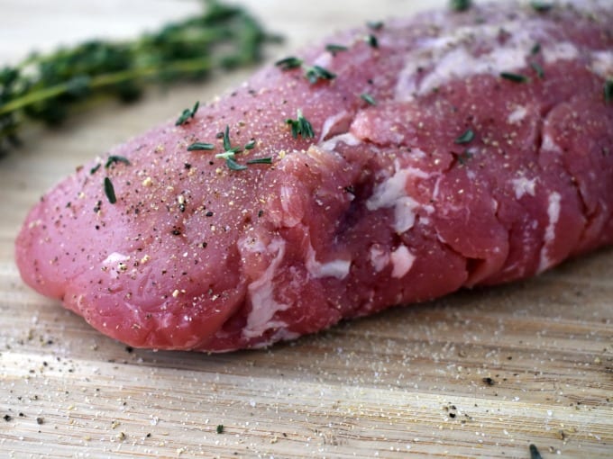 Uncooked pork tenderloin seasoned with salt, pepper and fresh thyme rests on a cutting board.