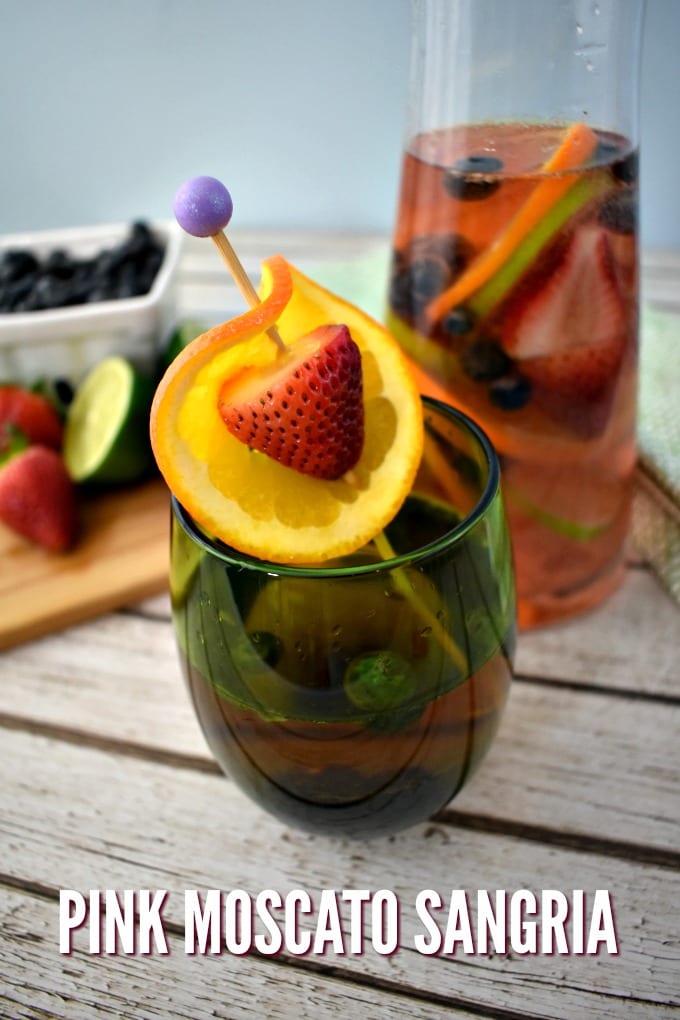 A stemless wine glass garnished with an orange slic and strawberry. A carafe of pink moscato sangria in the background next to a small cutting board with sliced limes and strawberries.