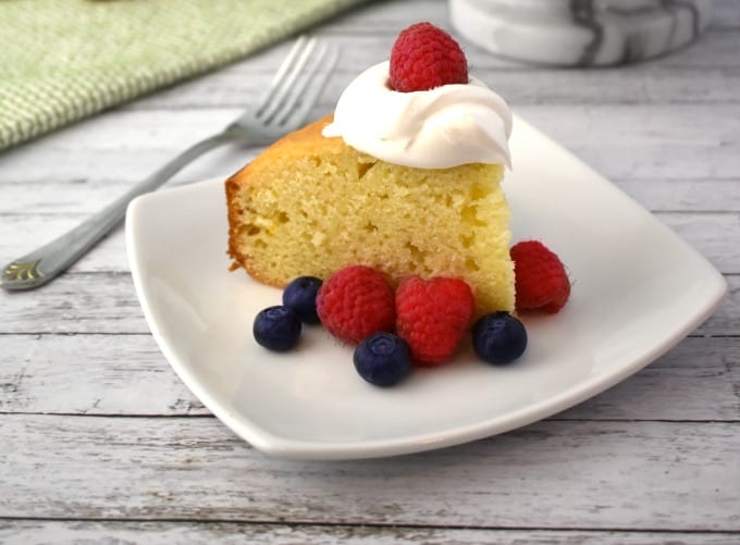 A slice of Yogurt Cake topped with a dollop of whipped cream and surrounded by fresh raspberries and blueberries sits on a white plate. A green and white hand towel can be seen in the background.