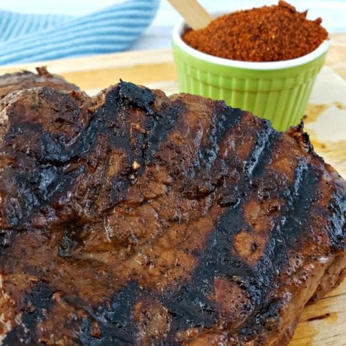 Grilled Petite Sirloin Steak on a cutting board. Int he background, a blue towel and a small green ramekin with Cowboy Steak Rub with a wooden spoon.