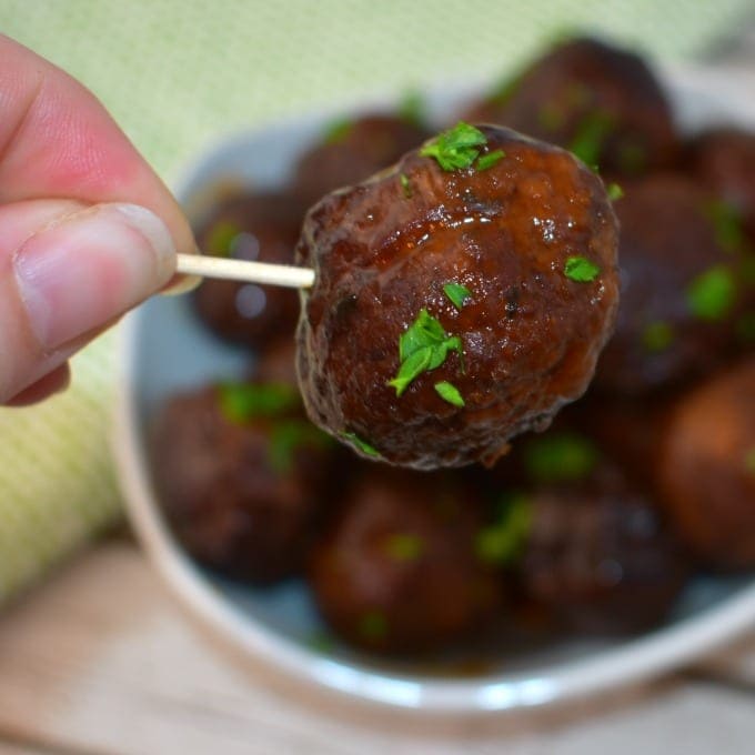 A hand holding a glazed meatball on a toothpick above a plate of meatballs, garnished with chopped parsley.