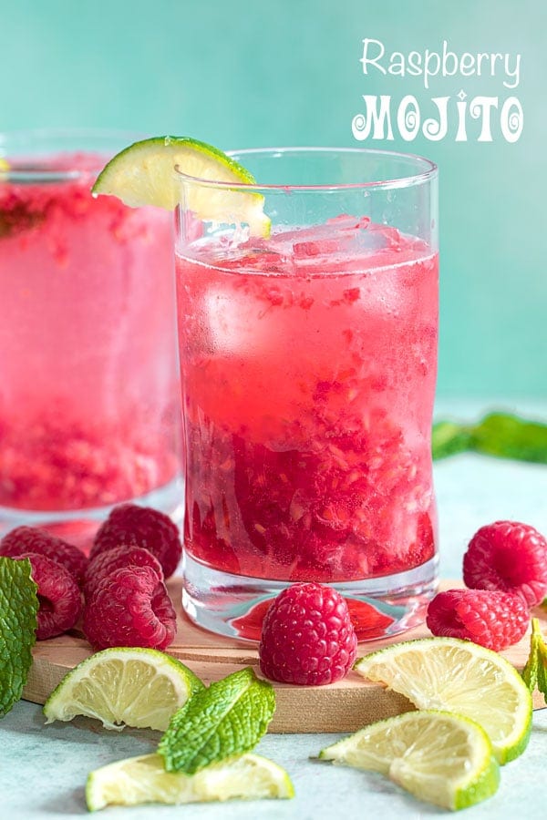 Front view of two glasses filled with raspberry mojito and surrounded by raspberries, lime and mint leaves