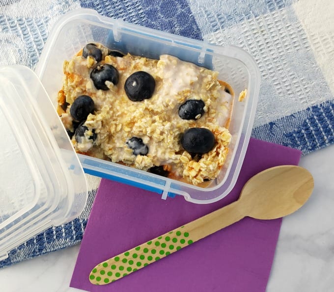 Blueberry Overnight Oats in a small containers on a marble counter with a purple napkin and wooden spoon.