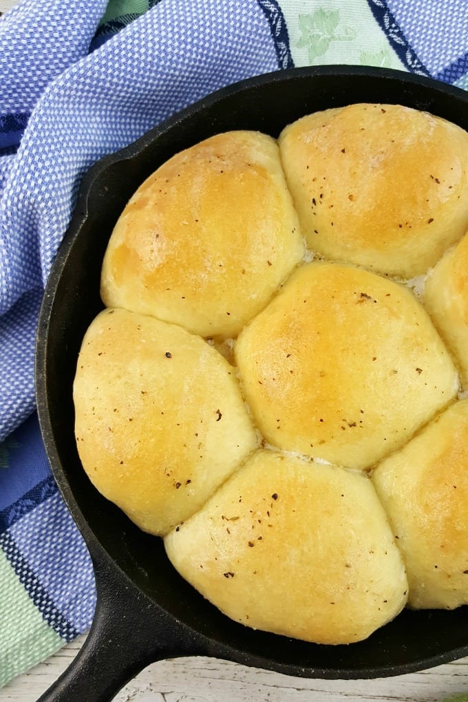 cast iron skillet with buttery dinner rolls next to a blue and green kitchen towel.