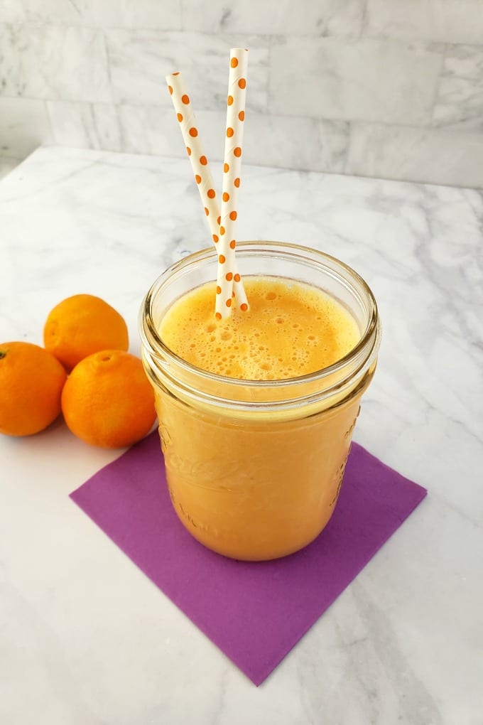 A clementine smoothie in a mason jar  with two orange polka dot paper straws next to fresh clementines.