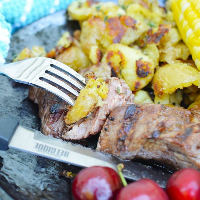 Close up of a fork with a bite of grilled sirloin tip next to fresh cherries, grilled potatoes and steamed corn on the cob.