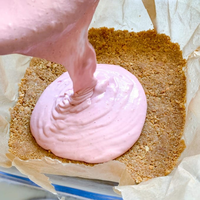 Pouring the cheesecake mixture into a lined baking pan holding a graham cracker crust.