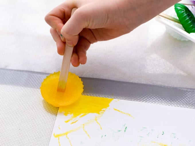 Ice Cube Painting - close up of a child holding a craft stick with frozen yellow ice cube to paint on paper.