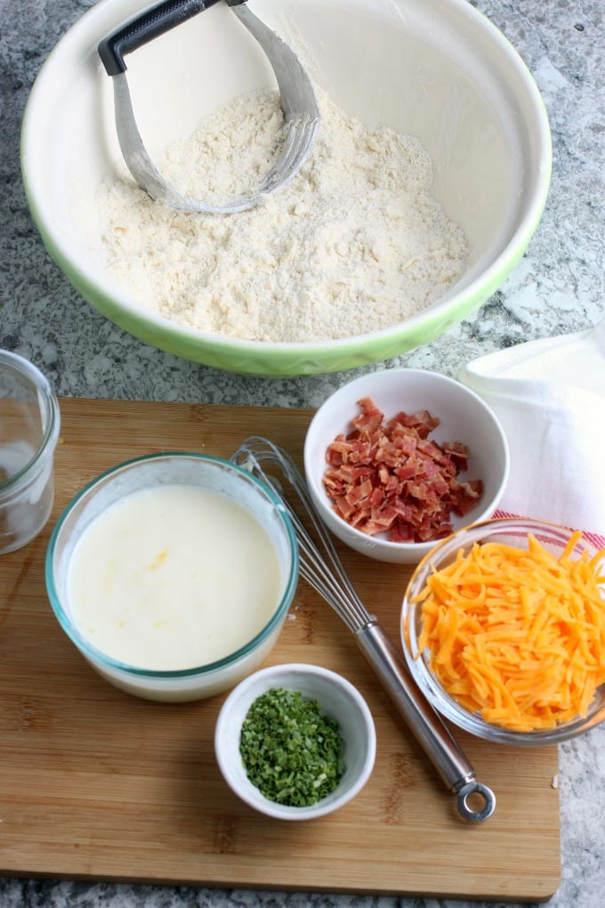 Four bowls containing buttermilk, bacon, cheese and chives on a wooden cutting board. Also pictured is a small whisk and a large mixing bowl full of flour and a pastry cutter. 
