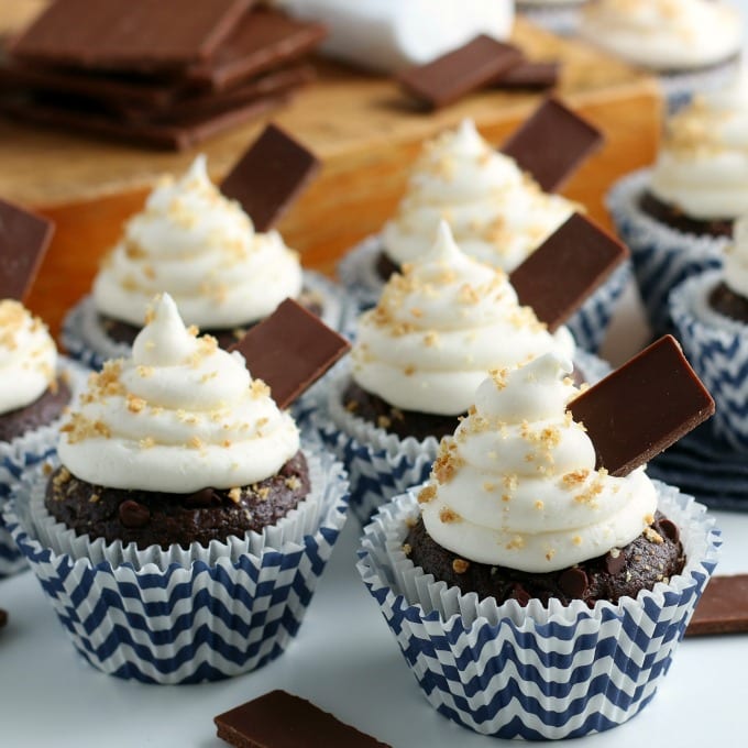 S'mores cupcakes in blue striped cupcake liners topped with marshmallow creme frosting, graham cracker crumbs and chunks of chocolate.