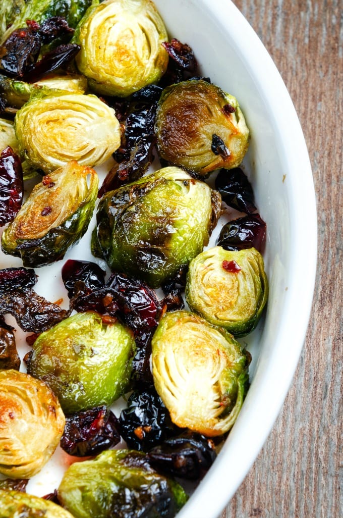 Roasted halved Brussels sprouts  and carnberries in a white casserole dish on a wood table. 
