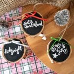 A birds-eye-view of wood slice Christmas ornaments, black with white text. Sitting on red and green plaid background.