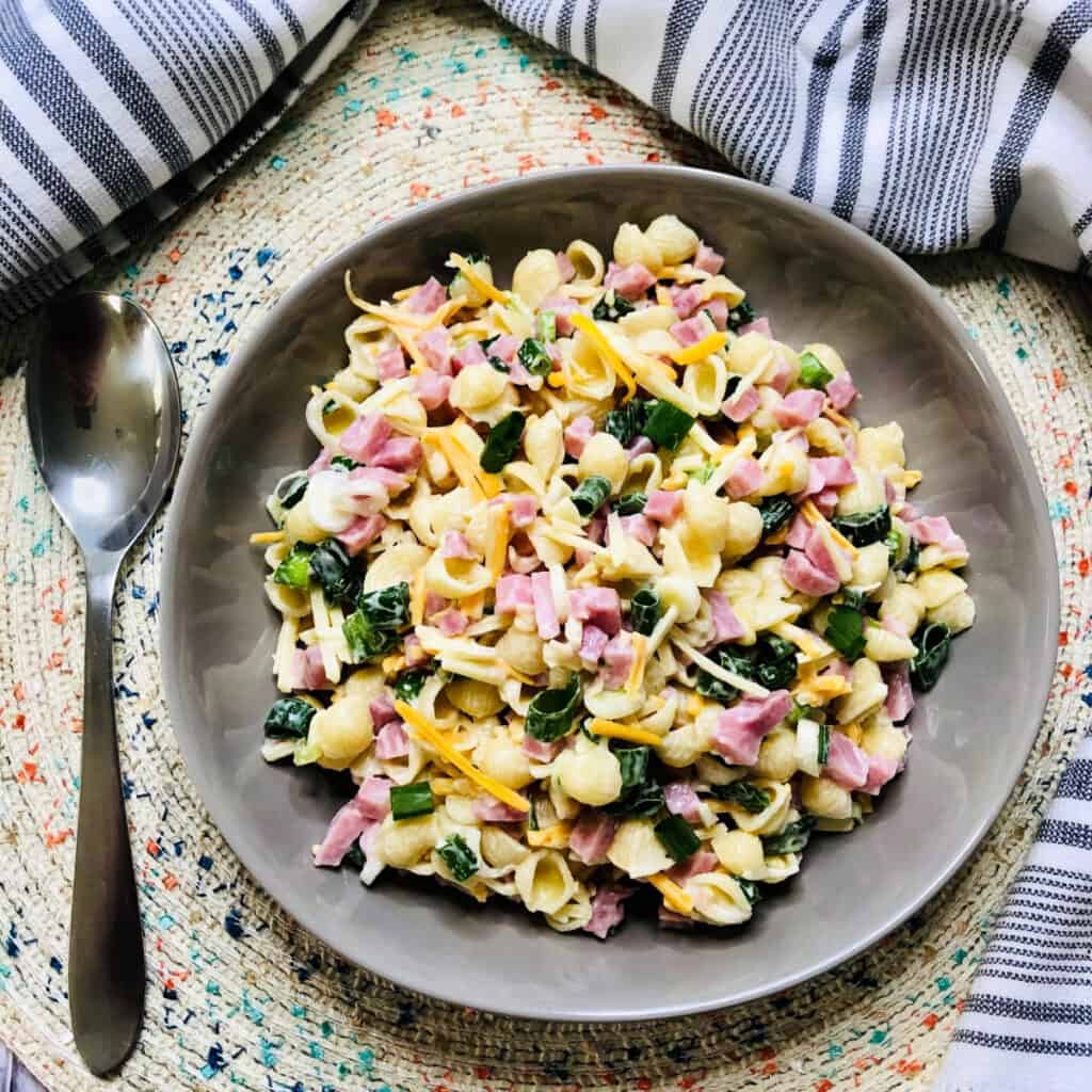 A gray bowl filled with Ham and Cheese Pasta Salad—shell pasta mixed with chopped ham, shredded cheese, and leafy greens—sits on a woven placemat next to a spoon and striped napkins.