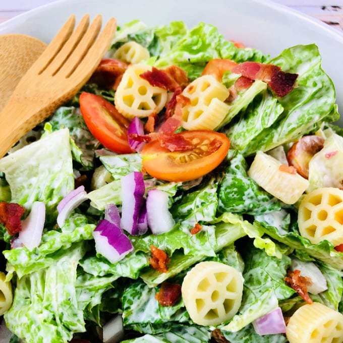 Easy Cold Dinner Ideas: a close-up of a fresh BLT Pasta Salad Recipe with romaine lettuce, grape tomato halves, red onion, crispy bacon pieces, and wheel-shaped pasta tossed in creamy dressing. A wooden fork and spoon rest on the side of the bowl.
