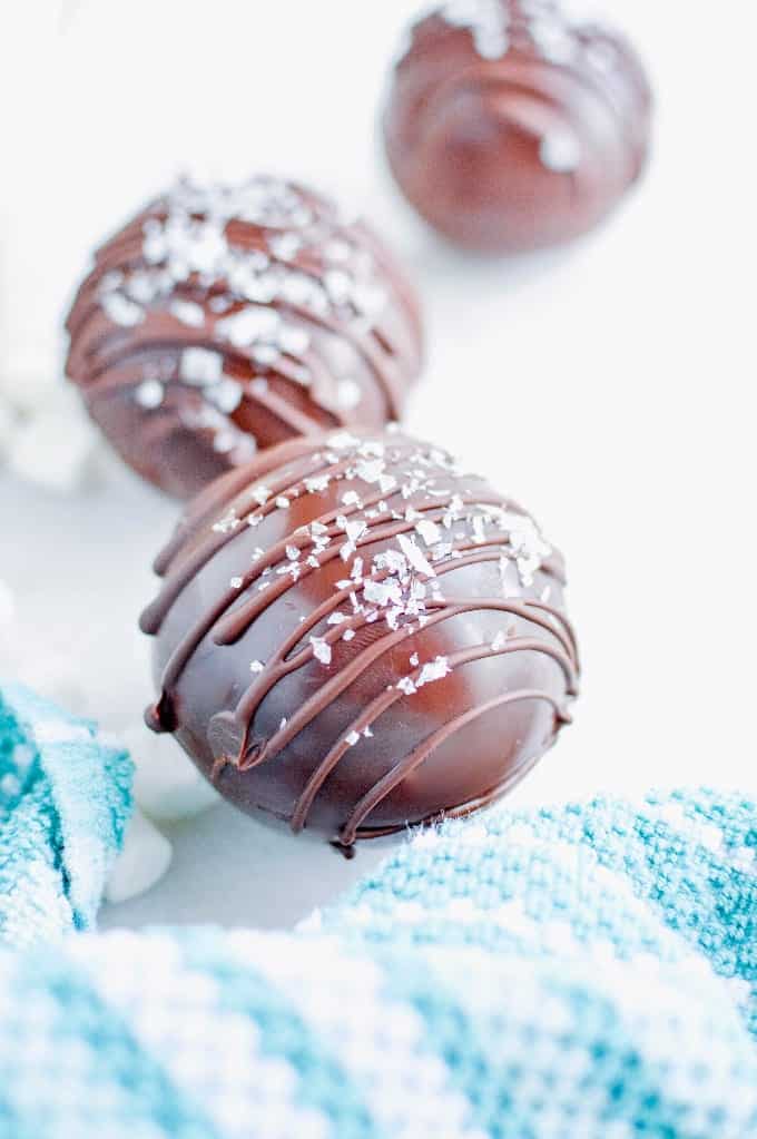 Close up image of salted caramel hot cocoa bombs topped with a chocolate drizzle and flaky sea salt next to a white and blue striped kitchen towel.