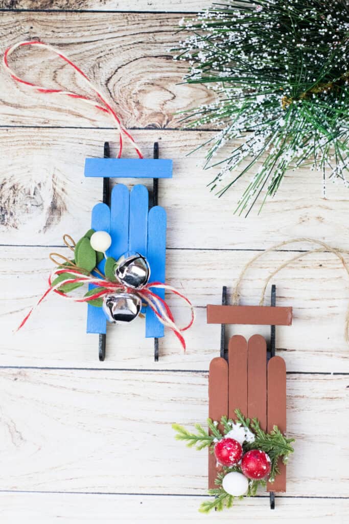 Two popsicle stick sled ornaments on a wood surface, surrounded by greenery.