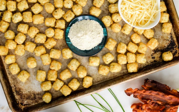 Tater tots and cheese on a sheet pan next to bacon strips.

