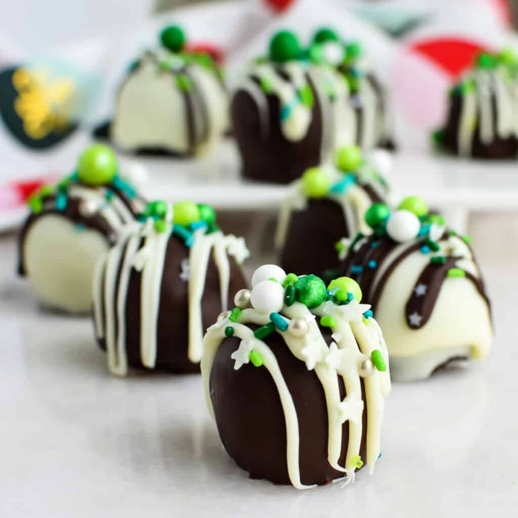 Chocolate truffles coated in dark and white chocolate, drizzled with white chocolate, and topped with green, white, and blue sprinkles—these football party sweets are finished with candy pearls and displayed on a white surface with a festive background.