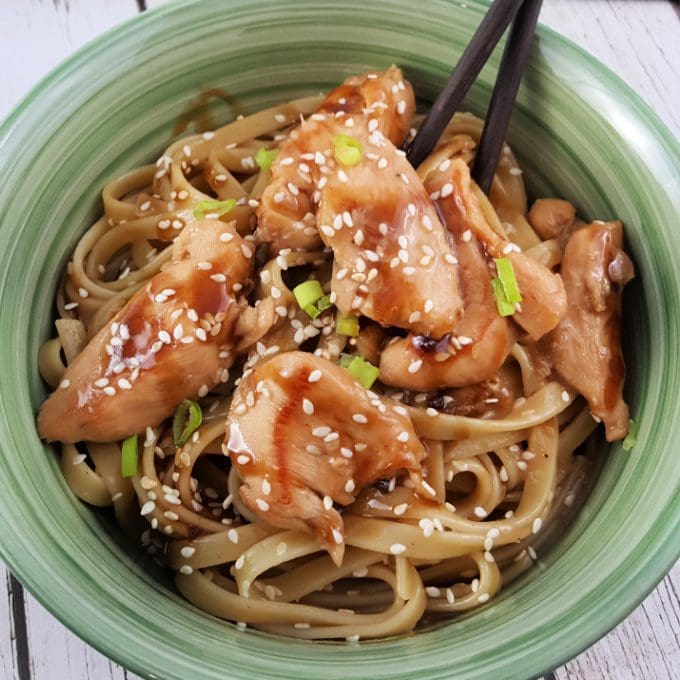 A green bowl filled with noodles topped with glazed chicken slices, sesame seeds, and chopped green onions; chopsticks are picking up a piece of chicken.