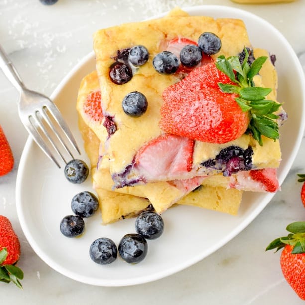 A plate with stacked square pieces of fluffy pancake topped with fresh strawberries and blueberries, with a fork and extra berries scattered around on a marble surface.