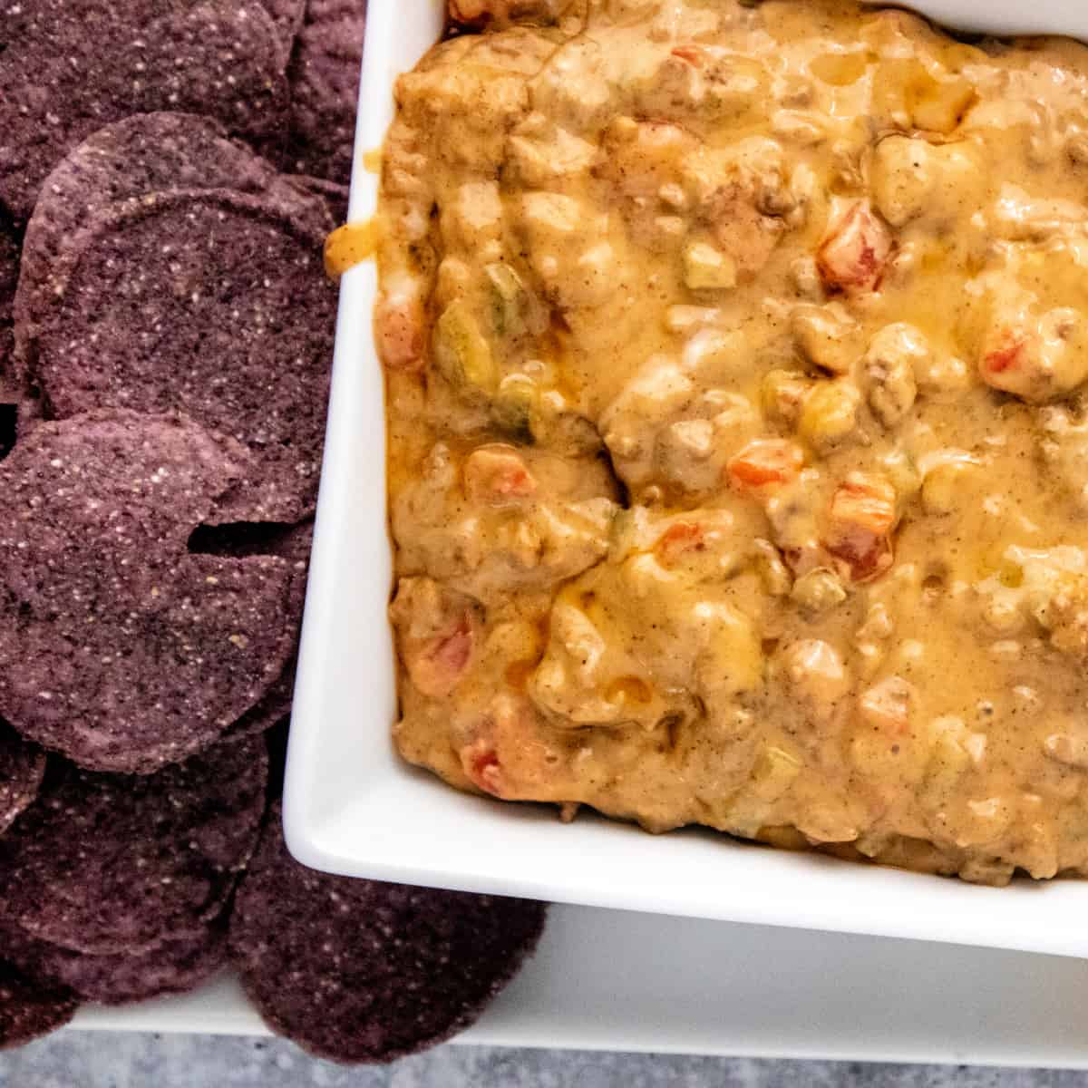 Close up of Beef Queso Dip in a square bowl next to blue corn tortilla chips.