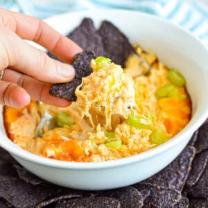 Close up of a hand scooping Cheesy Buffalo Chicken Dip with blue corn chips.