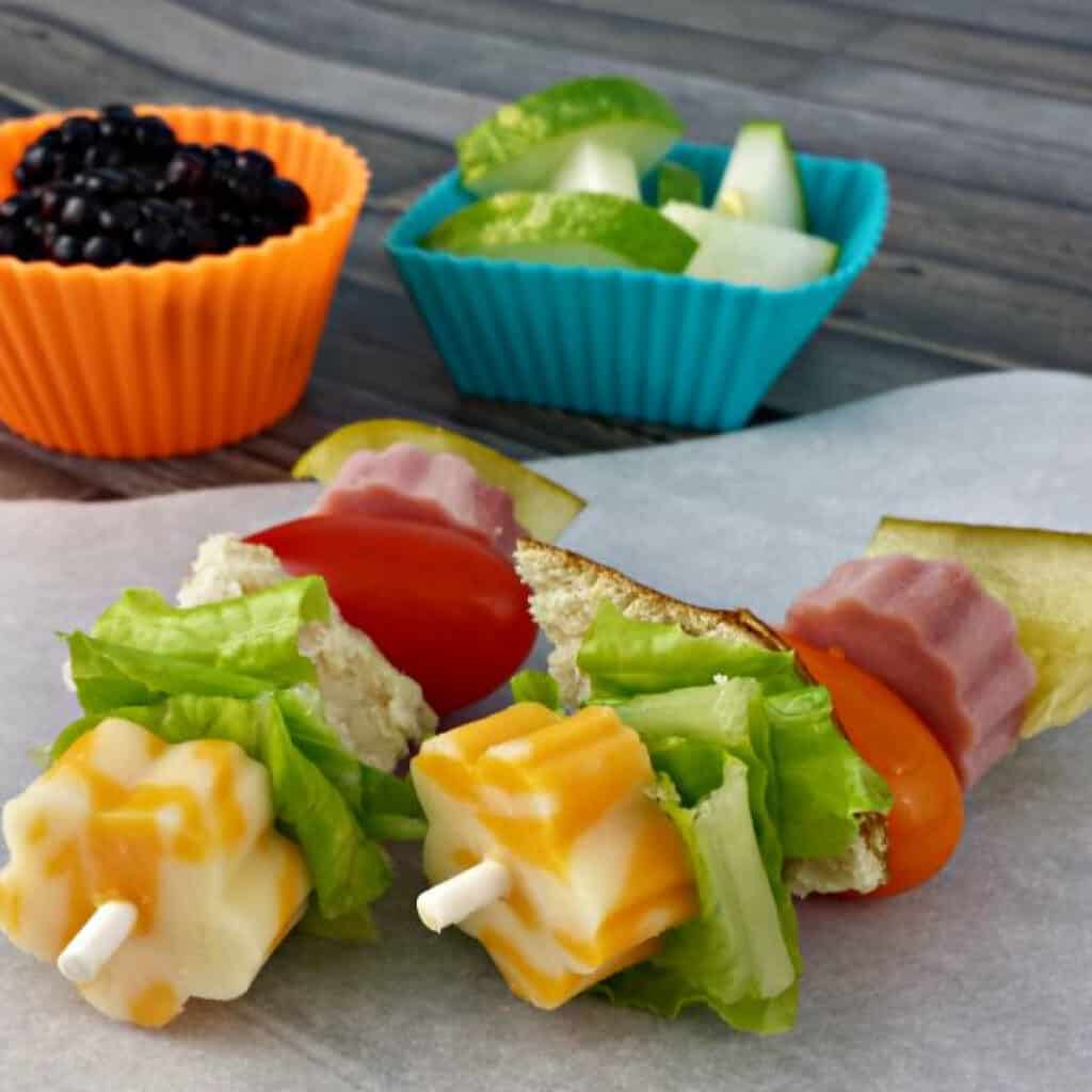No cook Dinner ideas: Two colorful skewers feature cubes of marbled cheese, lettuce, cherry tomato, lunch meat, and bread on parchment. Behind them, silicone cups hold blackberries and green apple slices with cucumber—perfect alongside Rotisserie Chicken Salad.
