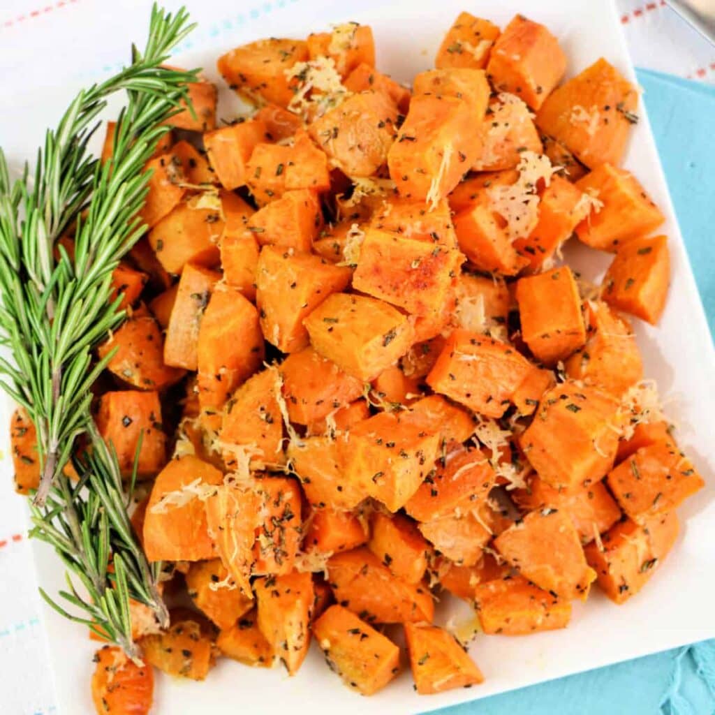 A square white plate piled with roasted sweet potato cubes topped with herbs and shredded cheese, garnished with fresh rosemary sprigs.