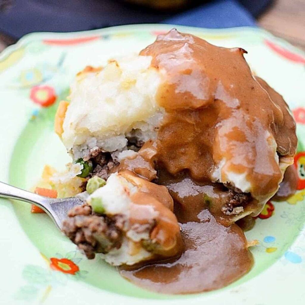 Ground beef recipes for dinner: a serving of shepherd’s pie with ground beef, peas, and mashed potatoes covered in brown gravy, on a floral plate. A fork has scooped up a portion from the side.