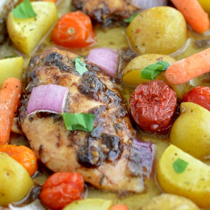 A close-up of baked chicken breast surrounded by baby potatoes, cherry tomatoes, carrots, red onion pieces, and herbs in a seasoned broth.