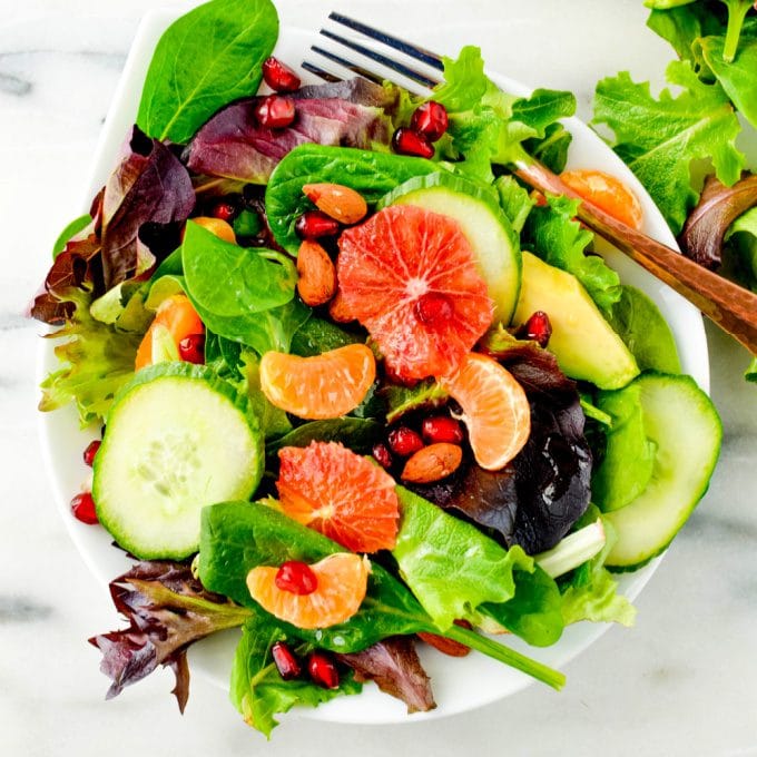 A salad with mixed greens, sliced cucumbers, avocado, orange segments, pomegranate seeds, and almonds in a white bowl with a fork.