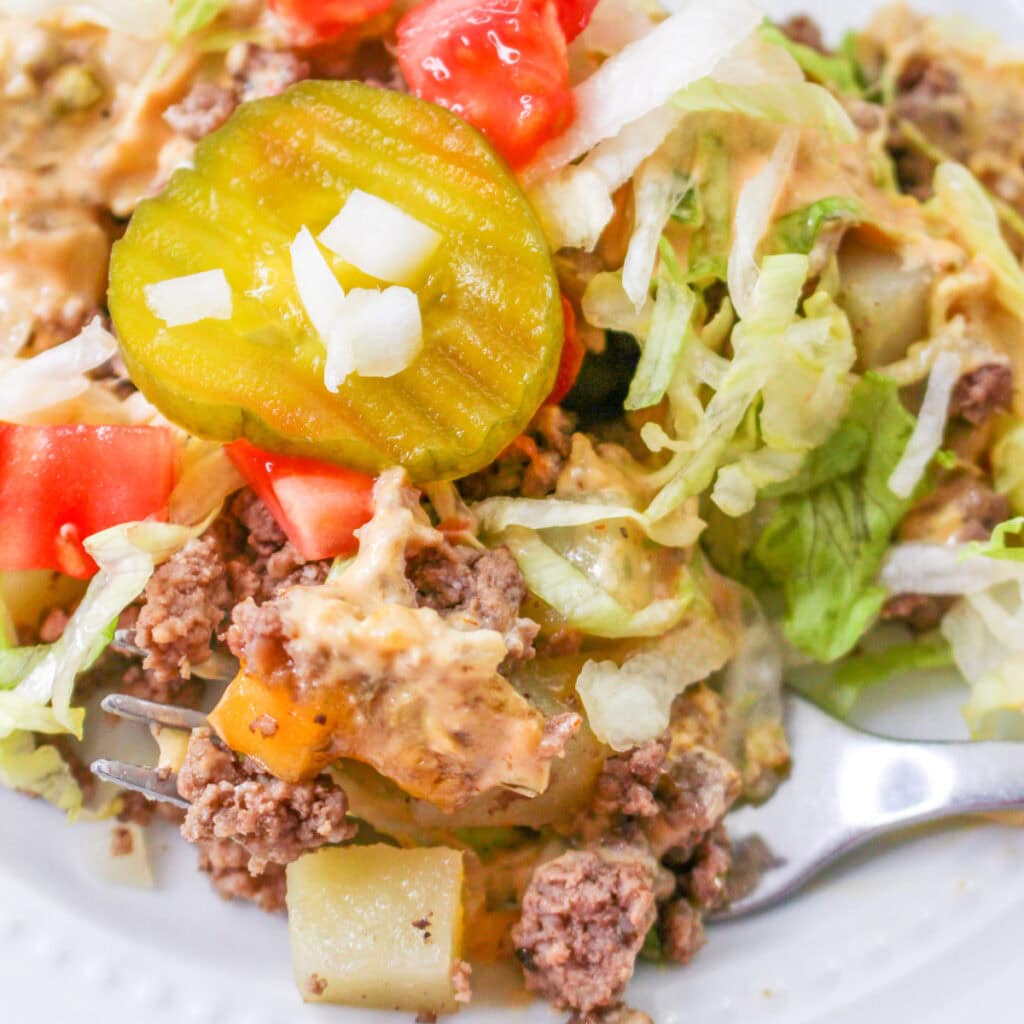 A close-up of a cheeseburger salad with ground beef, lettuce, diced tomatoes, cheddar cheese, pickles, onions, and a creamy sauce, served on a white plate with a fork.