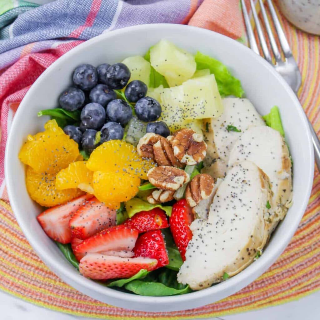 A white bowl filled with spinach, sliced chicken breast, strawberries, blueberries, orange segments, pineapple, pecans, and a sprinkle of poppy seeds, set on a colorful striped placemat with a napkin nearby.
