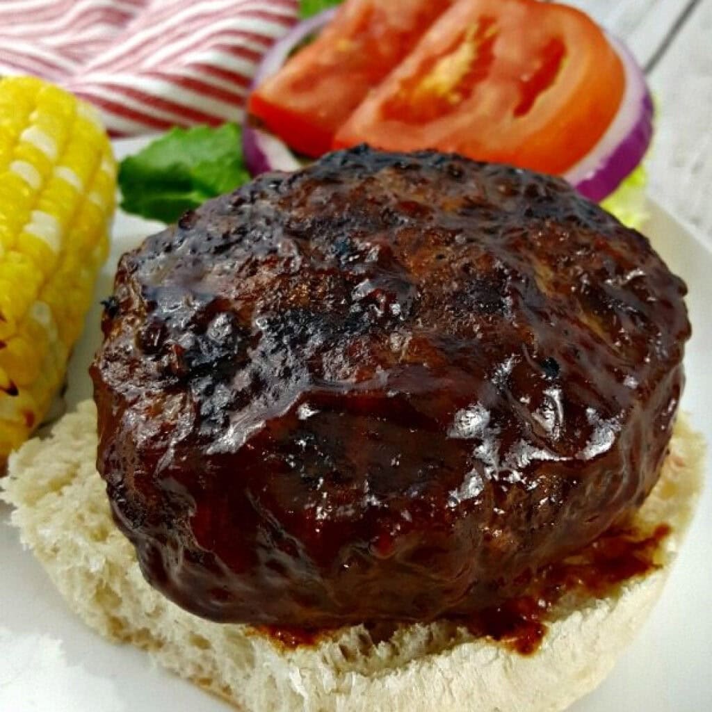 Close-up of a grilled hamburger patty covered in barbecue sauce on a bun, with corn on the cob and sliced tomato, lettuce, and onion in the background.