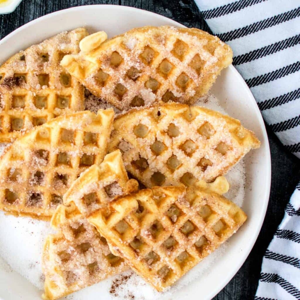 A white plate filled with golden-brown waffle pieces sprinkled generously with sugar, set on a dark surface next to a white and black striped cloth.