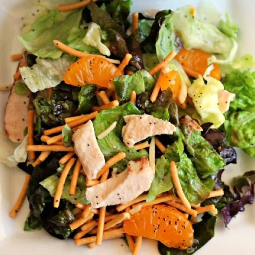 A salad with mixed greens, grilled chicken strips, mandarin orange slices, crunchy chow mein noodles, and poppy seed dressing on a white plate.