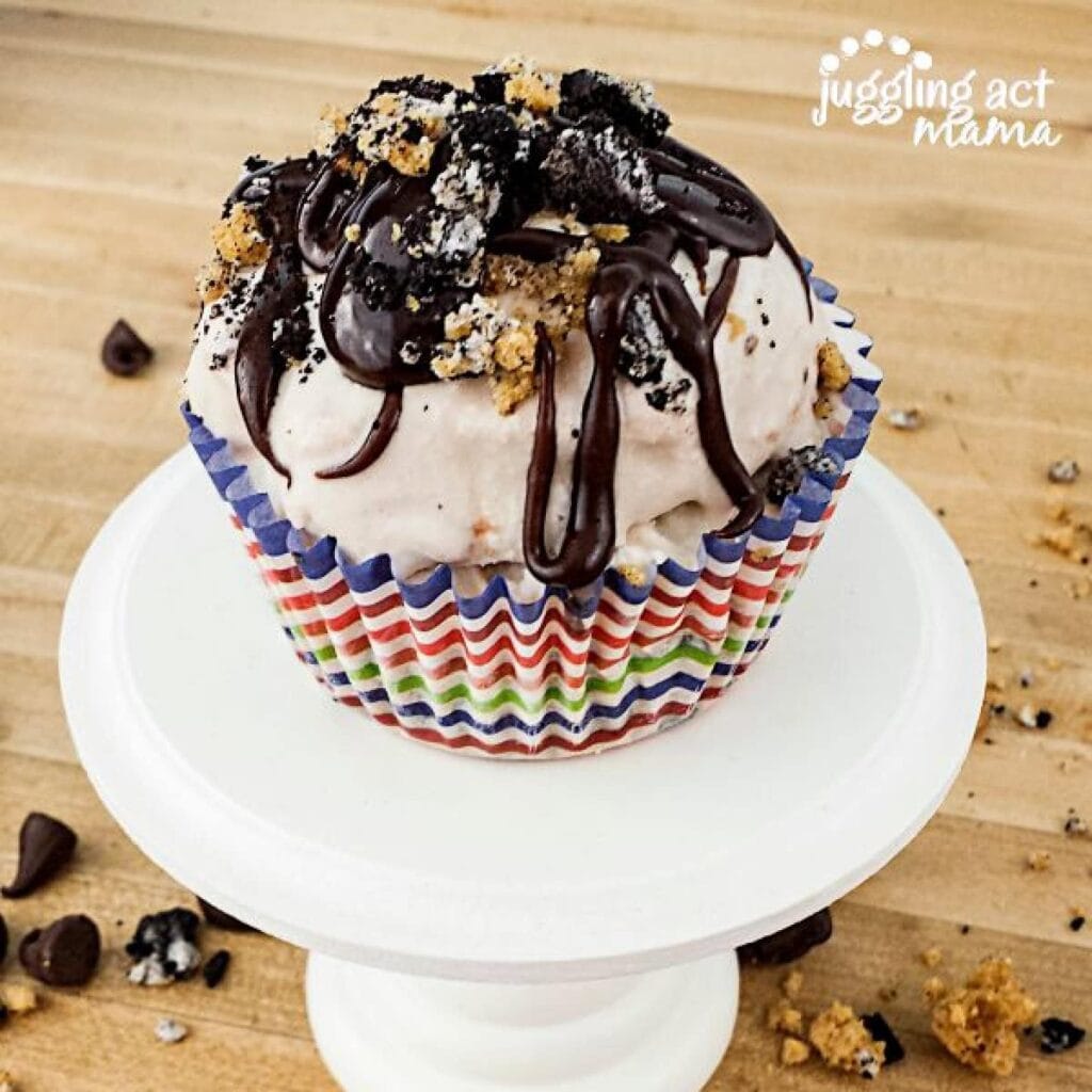 A cupcake with white frosting, chocolate drizzle, and cookie crumbs sits in a colorful striped wrapper on a white stand, with crumbs and chocolate chips scattered on a wooden surface.